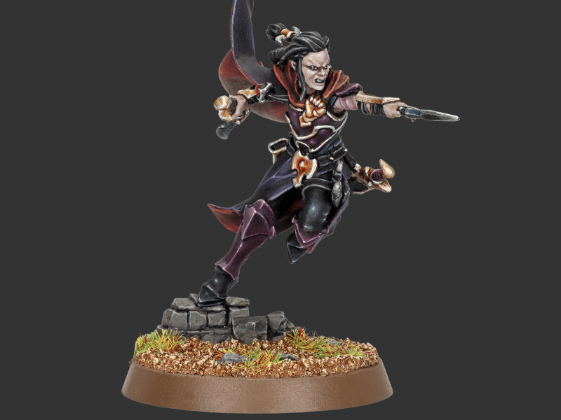 A painted fantasy miniature figurine of a female warrior in a dynamic leaping pose, holding a dagger, standing on a rocky base with grass and a brown circular platform against a dark gray background.