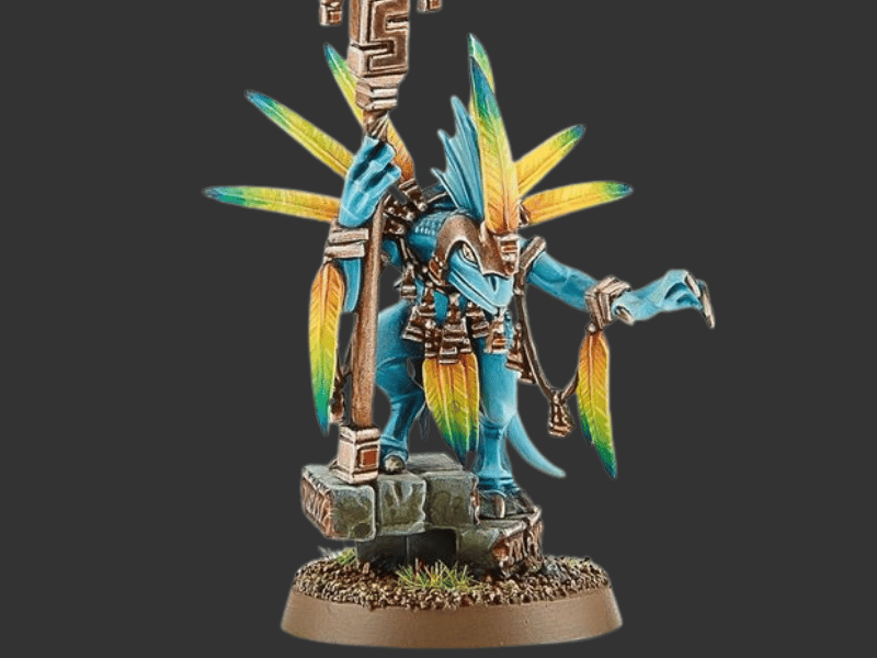 A painted fantasy miniature figurine of a blue-skinned deity or warrior standing on a stone base, with large yellow and teal feathered headdress-like wings, holding a staff, against a dark gray background.