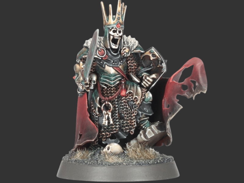 A painted fantasy miniature figurine of a skeletal crowned king or queen warrior holding a sword and shield, standing on a round base with a red tattered cape against a dark gray background.