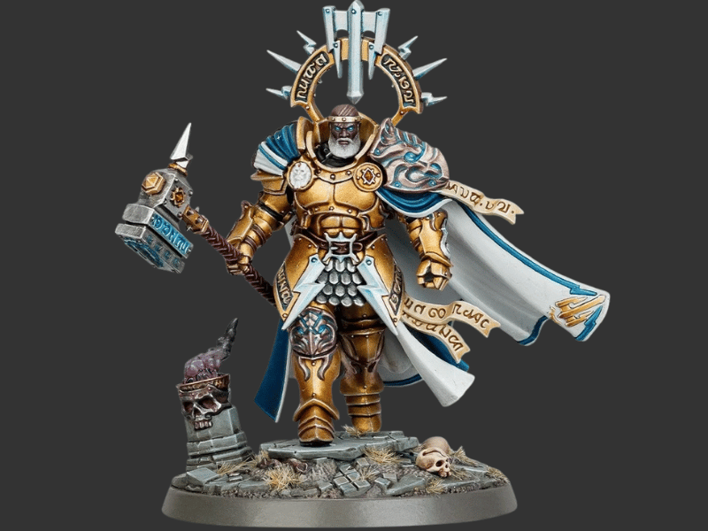 A highly detailed painted miniature figurine of a warrior in ornate gold armor with a flowing blue and white cape, holding a large hammer, standing on a rocky base against a dark gray background.