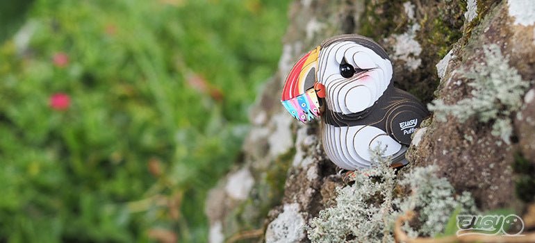 EUGY Puffin card model posed on a lichen-covered rock with grass and flowers behind