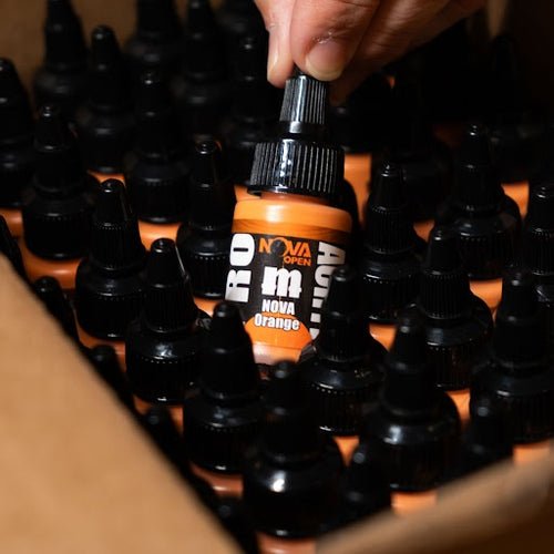 Hand lifting a Pro Acryl NOVA Orange 22ml bottle from a box of black-capped paint bottles