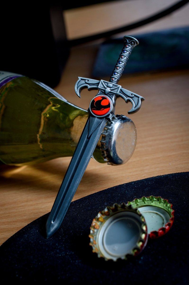 Sword of Omens bottle opener opening a bottle, with a bottle cap on the table