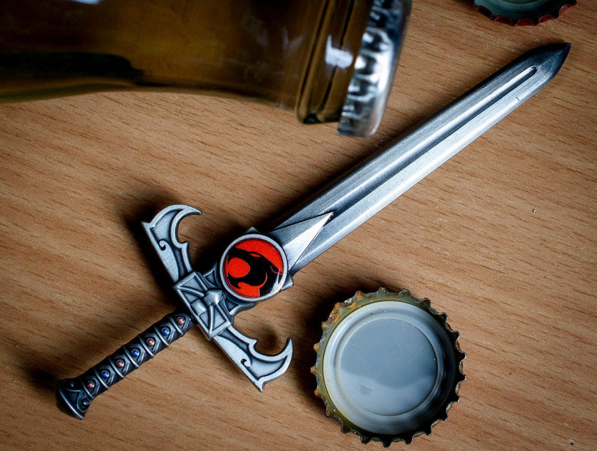 Sword of Omens bottle opener on a wooden table beside a bottle and bottle cap